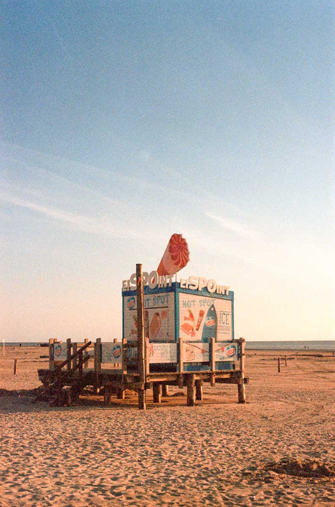 Ein Foto im Vintage-Stil von einem Eisstand an einem Sandstrand mit klarem Himmel, geschmückt mit verschiedenen bunten Werbeschildern.