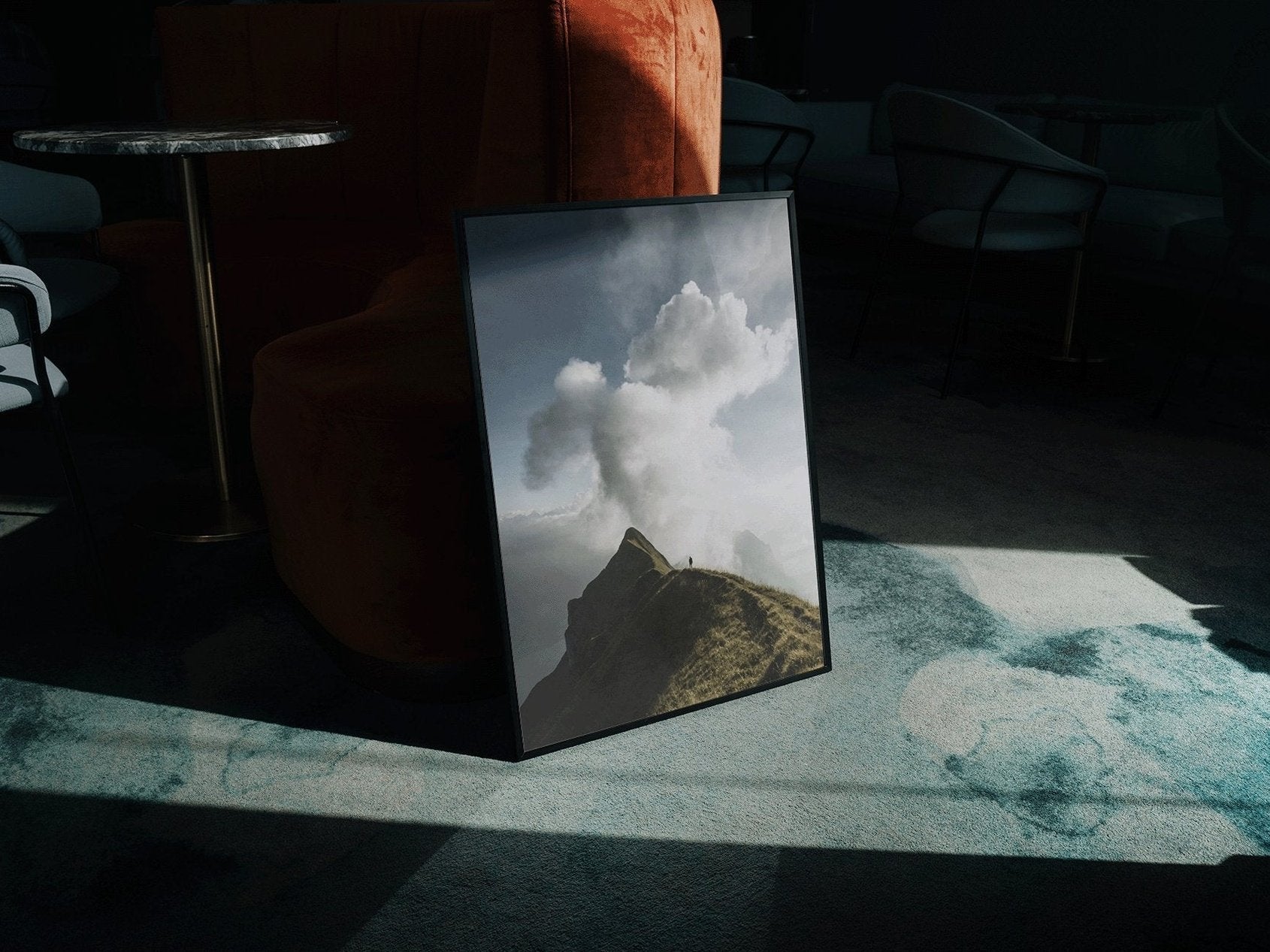 Gerahmtes Bild eines Berges mit Wolken, an einen orangefarbenen Stuhl gelehnt auf einem schwach beleuchteten, gemusterten Boden in einem Raum mit Stühlen und Tischen.