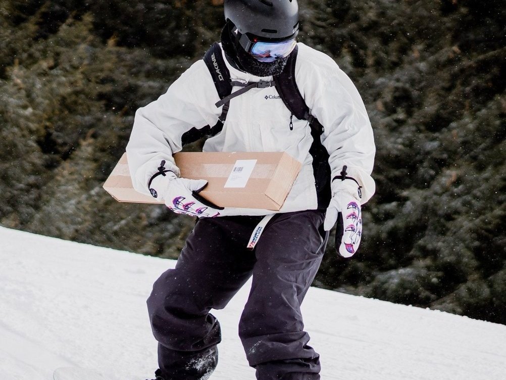 Eine Person in Winterkleidung und Helm trägt ein rechteckiges Paket, während sie mit dem Snowboard einen schneebedeckten Hang hinunterfährt.