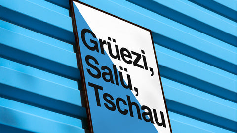 Ein Schild mit der Aufschrift „grüezi, salü, tschau“ in schwarzer Schrift auf weissem Grund, montiert vor einer blau gestreiften Fassade unter einem klaren Himmel.