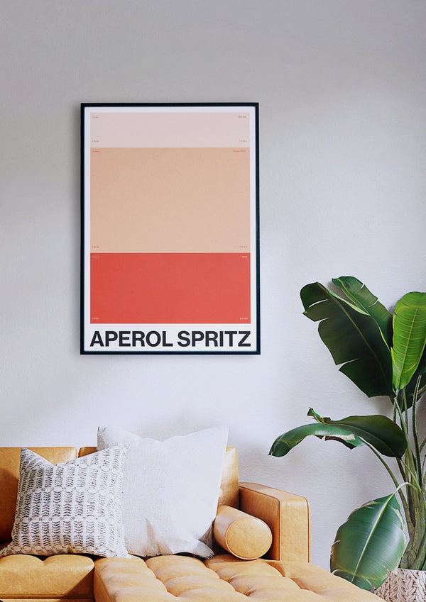 Ein gerahmtes „APEROL SPRITZ“-Poster mit minimalistischem Design hängt über einem senffarbenen Sofa, das mit zwei Sofakissen geschmückt ist, während rechts davon eine große grüne Pflanze steht.