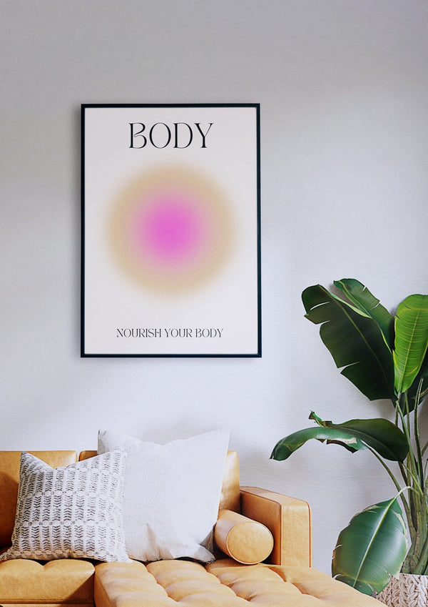 Ein gerahmtes Poster mit dem Titel „BODY“ und der Aufschrift „NOURISH YOUR BODY“ hängt über einem senffarbenen Sofa mit weißen Kissen, neben einer grünen Pflanze vor einer weißen Wand.