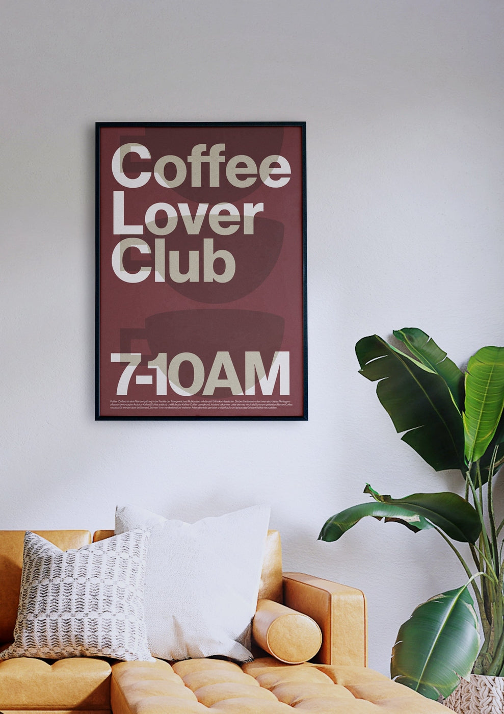 Ein gerahmtes „Coffee Lover Club“-Poster hängt an einer Wand über einem hellbraunen Sofa mit weißen und gemusterten Kissen, das neben einer großen grünen Pflanze steht.