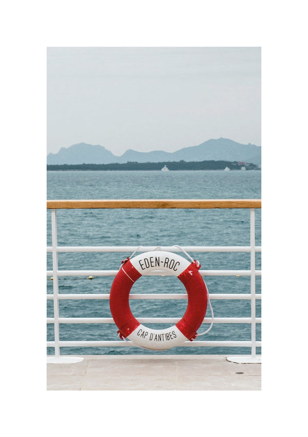 Ein Rettungsring von Eden Roc mit einem rot-weißen Design und der Aufschrift „Eden-Roc Cap d'Antibes“ hängt an einem Zaun mit einem Gewässer und entfernten Bergen im Hintergrund.