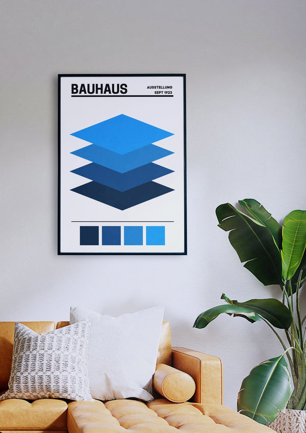 Ein gerahmtes „Bauhaus Layers 1“-Poster mit geometrischen blauen Formen hängt an einer weißen Wand über einem senfgelben Sofa mit weißen und grauen Kissen. Rechts neben dem Sofa steht eine grüne Pflanze.