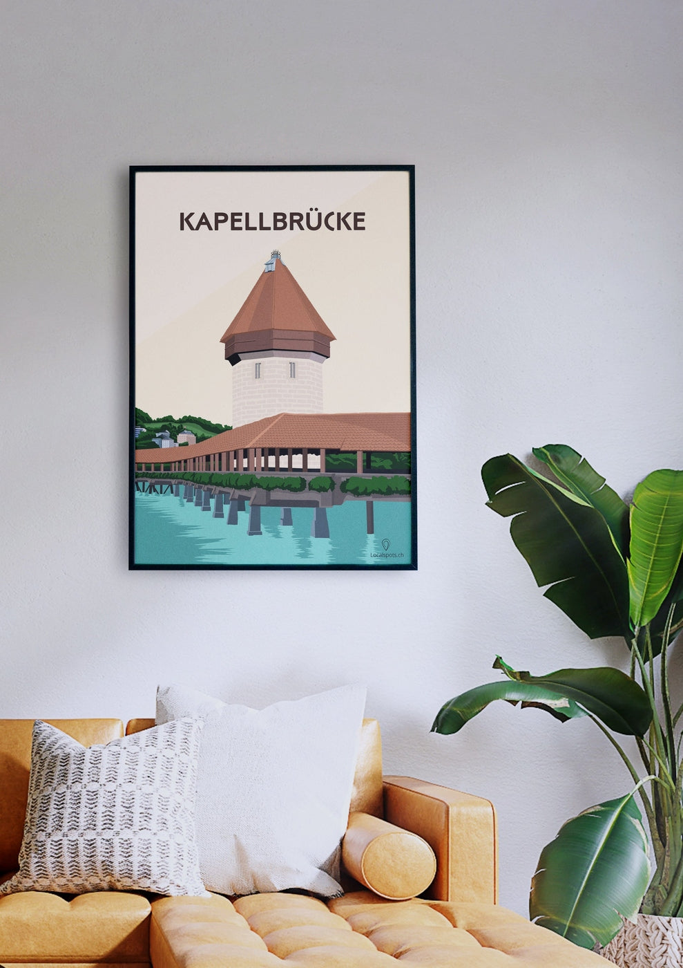 Ein gerahmtes Poster mit dem Titel „Kapellbrücke“, das eine Holzbrücke mit Turm zeigt, hängt an einer weißen Wand über einem senffarbenen Sofa mit weißen und gemusterten Kissen. Neben dem Sofa steht eine grüne Pflanze.