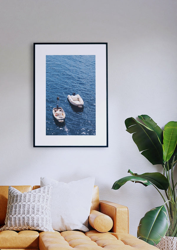 Ein gerahmtes Foto mit dem Titel „Mediterrane Meditation“, das zwei Boote auf dem Meer zeigt, hängt an einer weißen Wand über einem senffarbenen Sofa mit weißen und gemusterten Kissen. Rechts neben dem Sofa steht eine große grüne Pflanze.