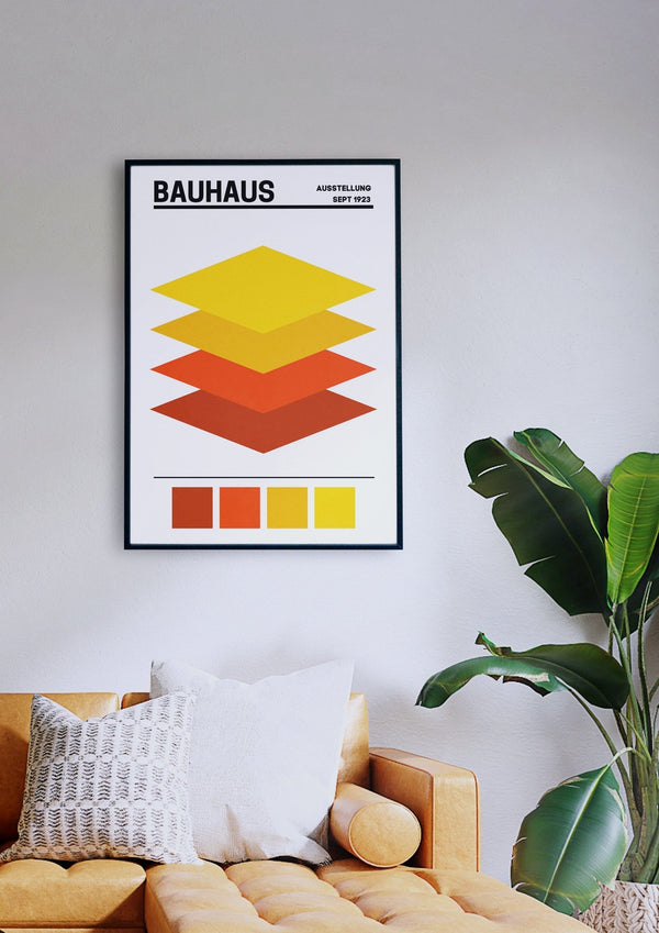 Ein gerahmter Kunstdruck „Bauhaus Layers 2“ mit geometrischen Schichten in Gelb- und Orangetönen hängt an einer weißen Wand über einem senffarbenen Sofa mit weißen und gemusterten Kissen. Daneben steht eine grüne Pflanze.