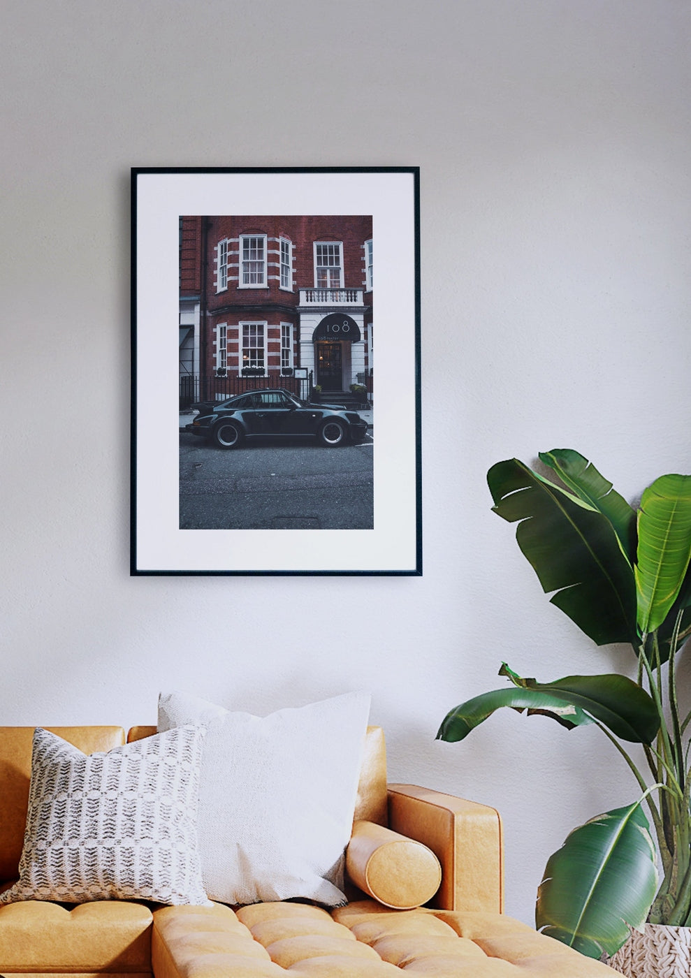 Das gerahmte Foto „Streets of London“, das einen schwarzen Oldtimer zeigt, der vor einem roten Backsteingebäude geparkt ist, hängt an einer weißen Wand über einem hellbraunen Sofa mit weißen und gemusterten Kissen. Neben dem Sofa steht eine grüne Pflanze.