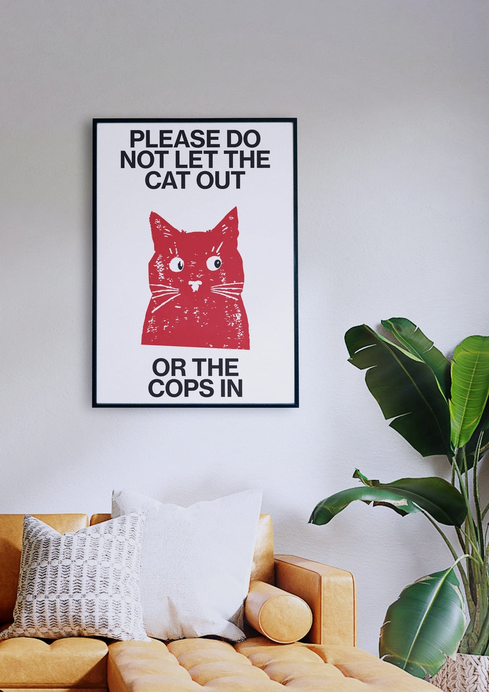 Ein gerahmtes „Sus Cat“-Poster mit einer roten Katzenillustration und dem Text „Bitte lassen Sie die Katze nicht raus oder die Polizei rein“ hängt an einer weißen Wand über einem senfgelben Sofa mit Kissen neben einer grünen Topfpflanze.