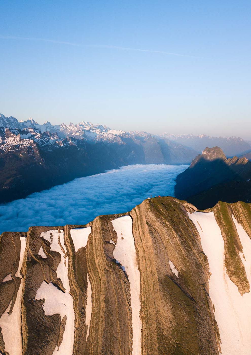 Eine Luftaufnahme eines schneebedeckten Gebirgskamms mit dem Nebelmeer im Hintergrund.