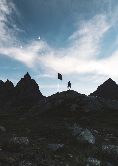 Eine Person steht zur Dämmerung auf dem Gipfel eines Berges mit einer Schillerlichtflagge.
